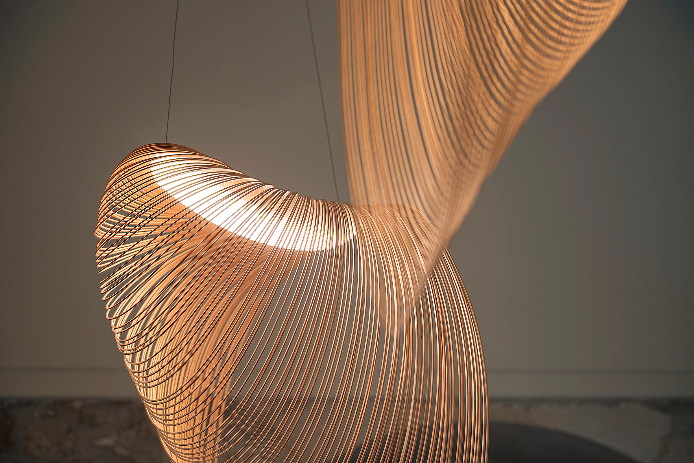 Le sottili lamelle curve di due lampade a sospensione illuminate, una a fuoco e l’altra sfocata in primo piano.