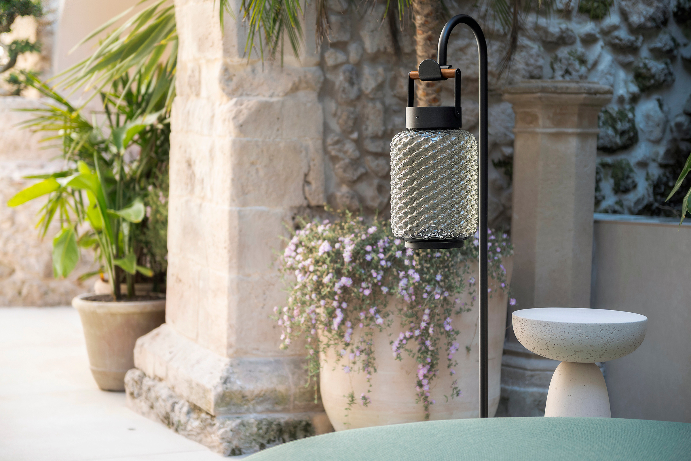 Una lampada portatile in vetro su un supporto nero, accanto a un tavolino chiaro, fiori e pilastri in pietra nella corte.
