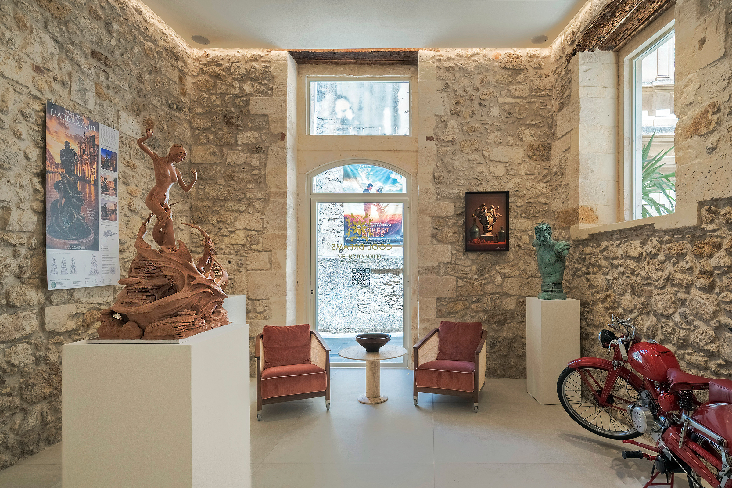 Due poltrone tra sculture, dipinti e una motocicletta d’epoca nella galleria affacciata sulla strada.