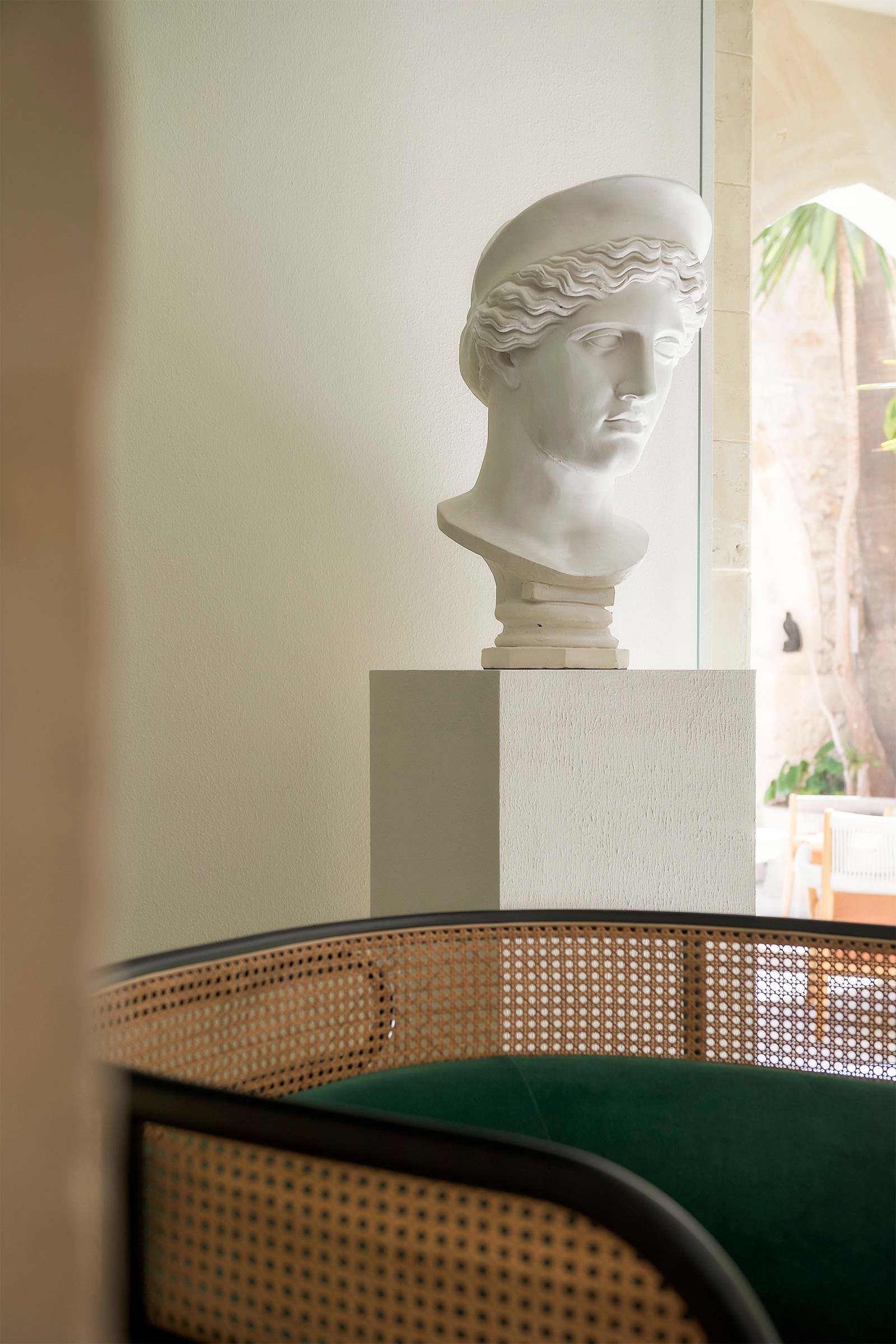 Un busto bianco accanto all’affaccio sulla corte, con una seduta in legno, midollino e tessuto verde in primo piano.