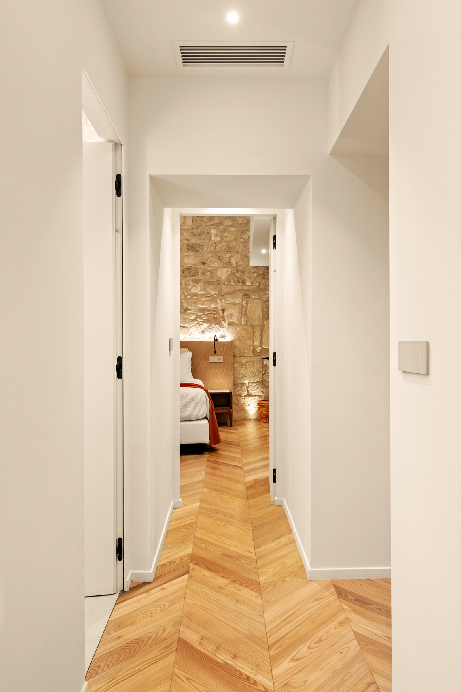 Una sequenza di soglie verso la camera, con griglia dell’aria a soffitto, parquet e un letto sullo sfondo davanti alla parete in pietra.