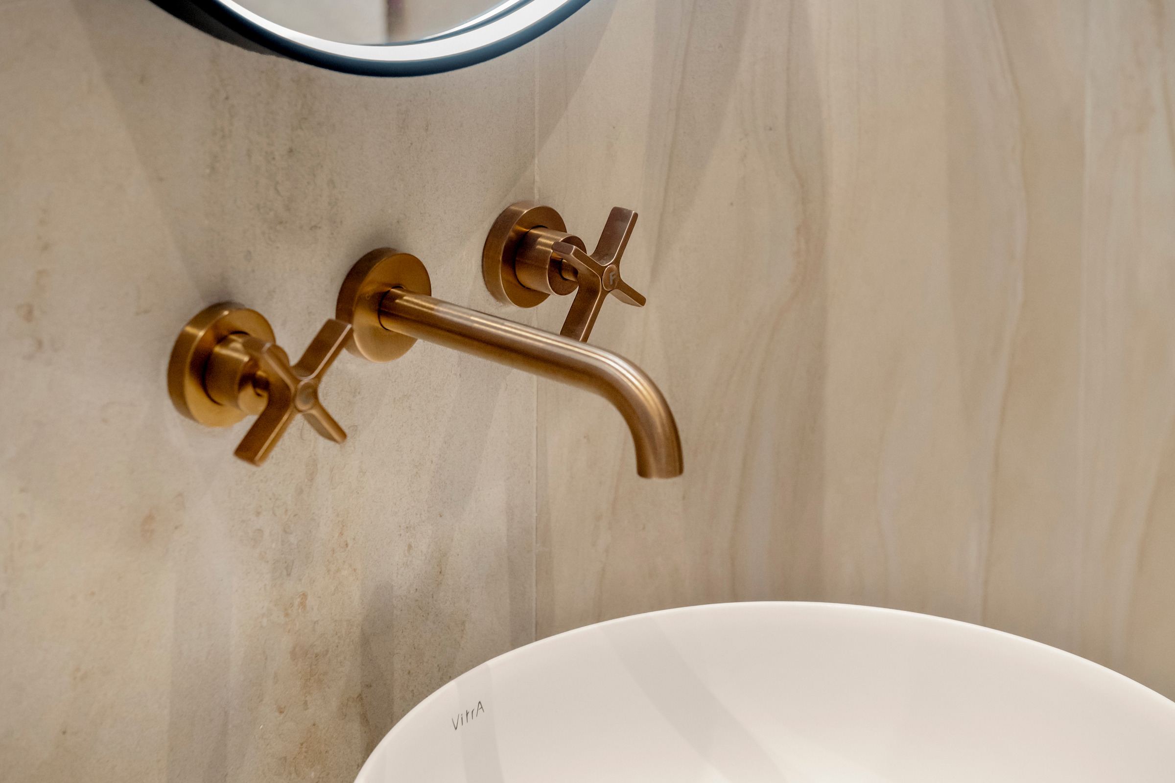 Rubinetteria a parete con finitura calda sopra un lavabo bianco, su un rivestimento dalle venature chiare.