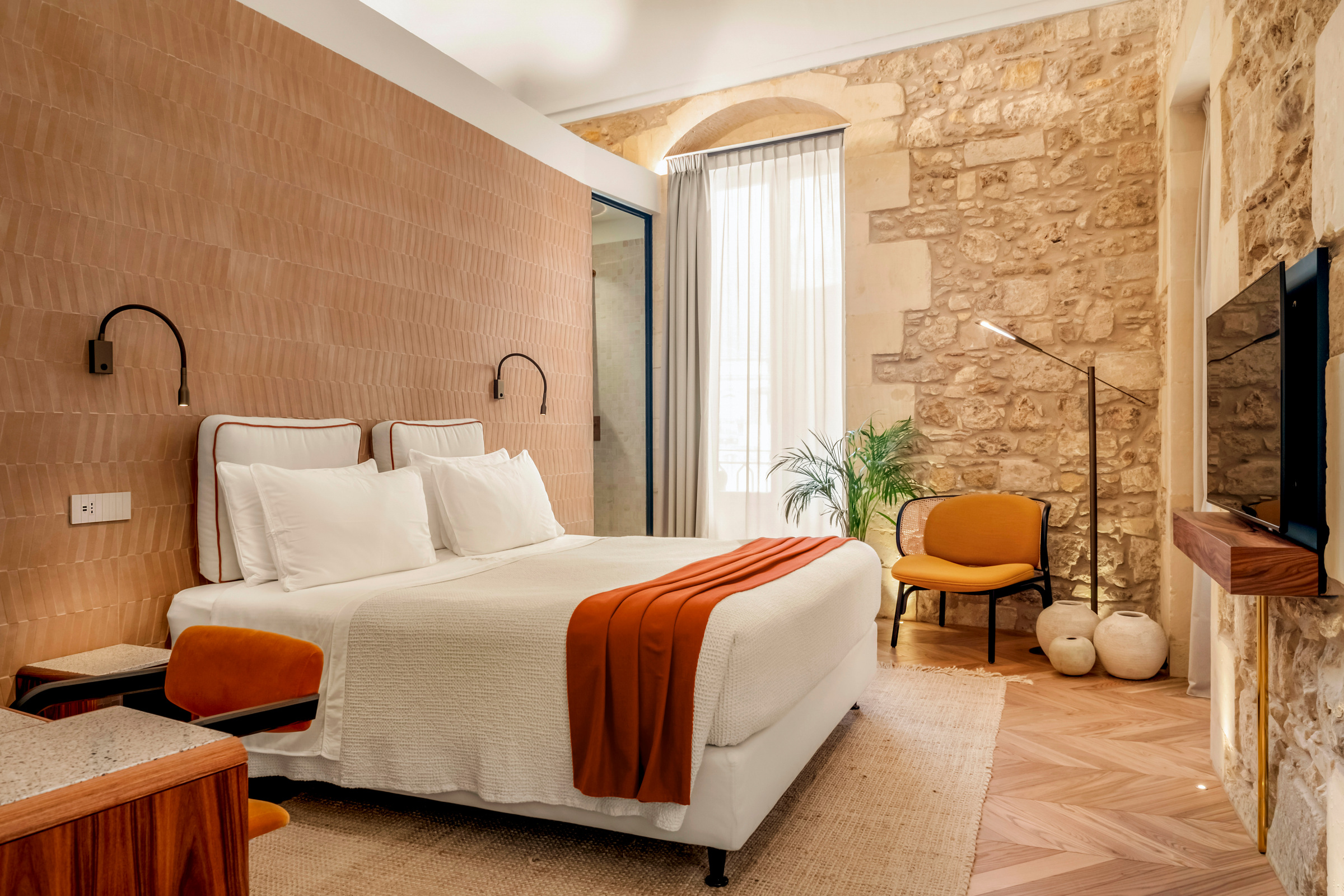 Letto con biancheria chiara, coperta terracotta e poltrona ocra accanto alla finestra.